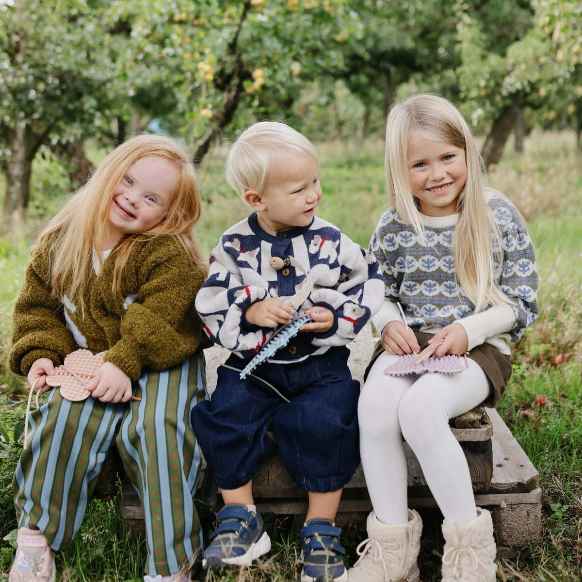 Three kids smile outdoors, playing with mushie’s Silicone Sensory Toy for fine motor fun.