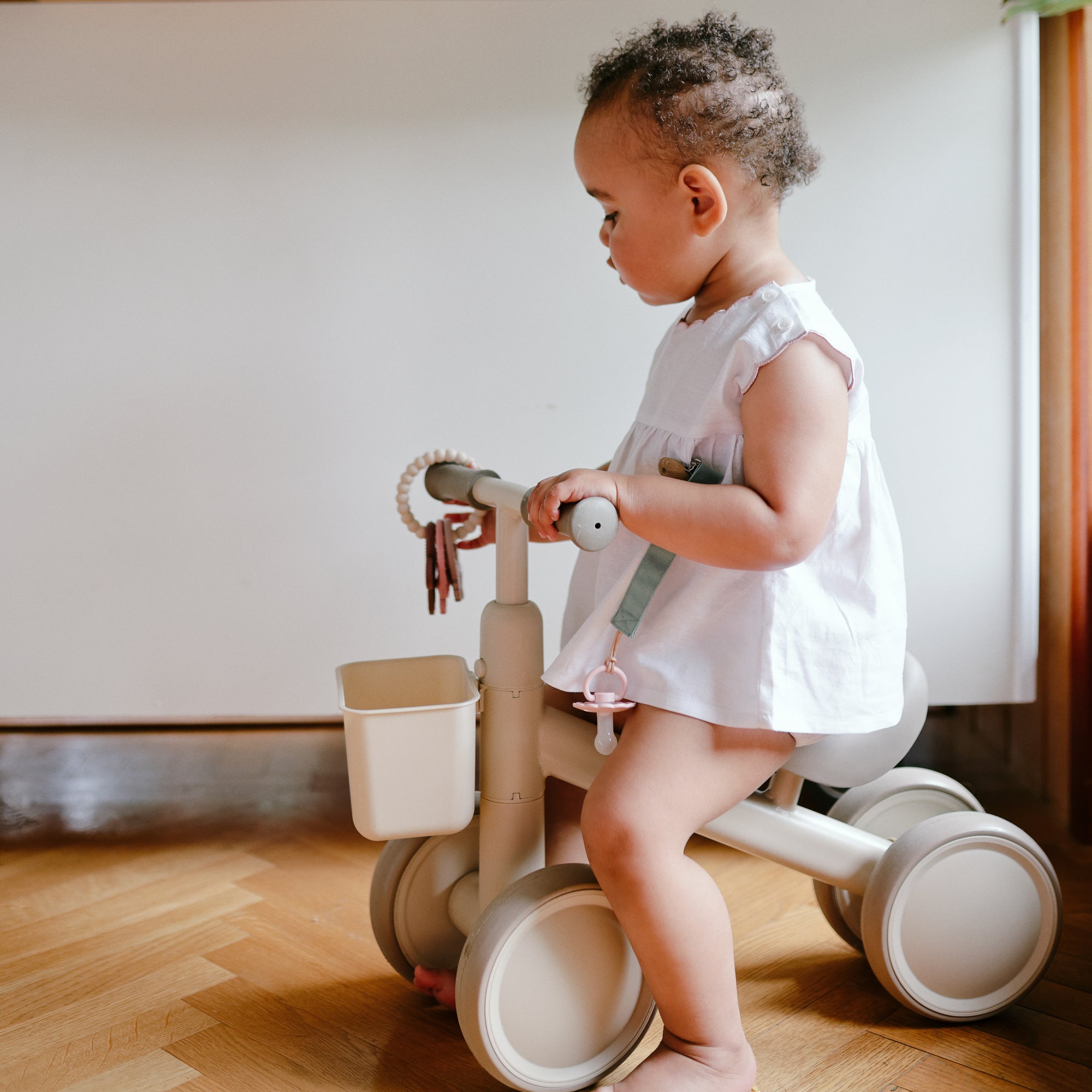 Toddler in white dress holding a mushie Teething Ring, sitting on a beige tricycle indoors.