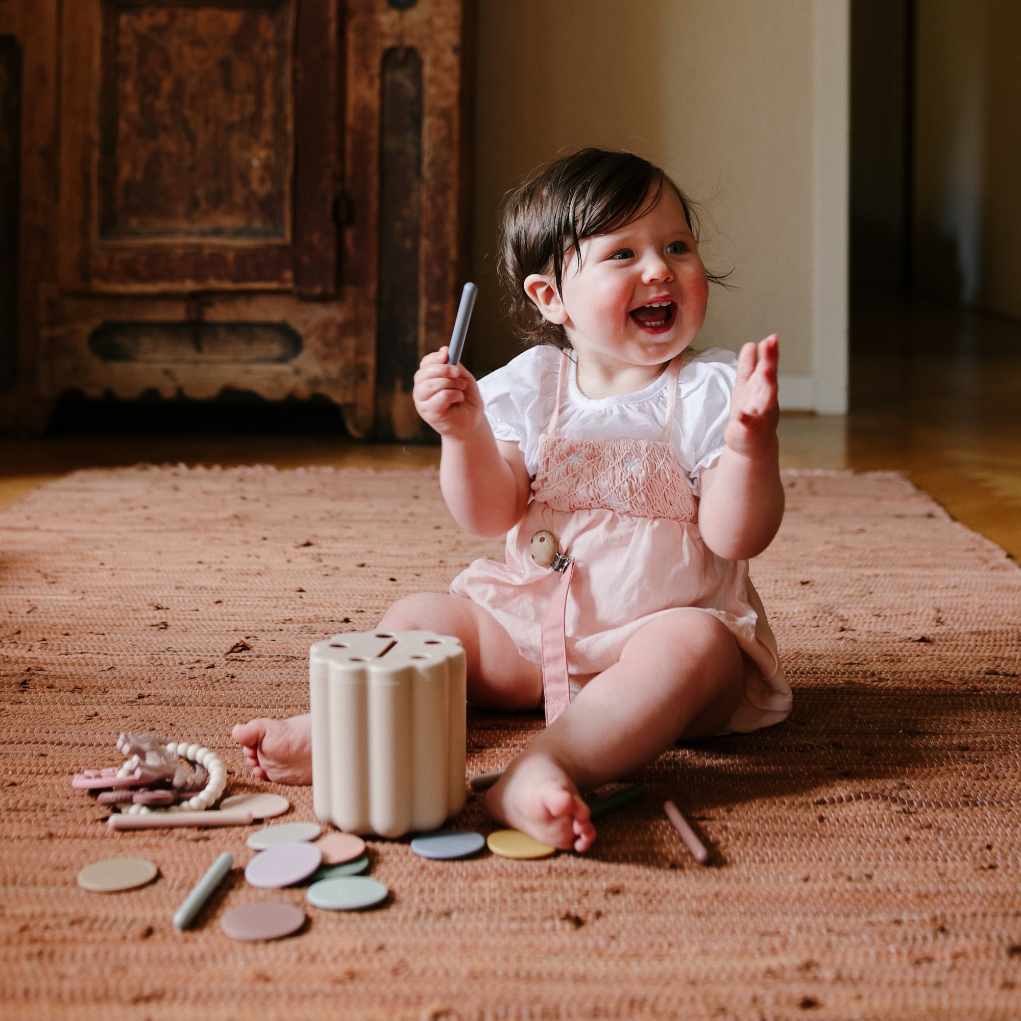 Smiling baby on a rug, playing with mushie Teething Ring and colored chalk.
