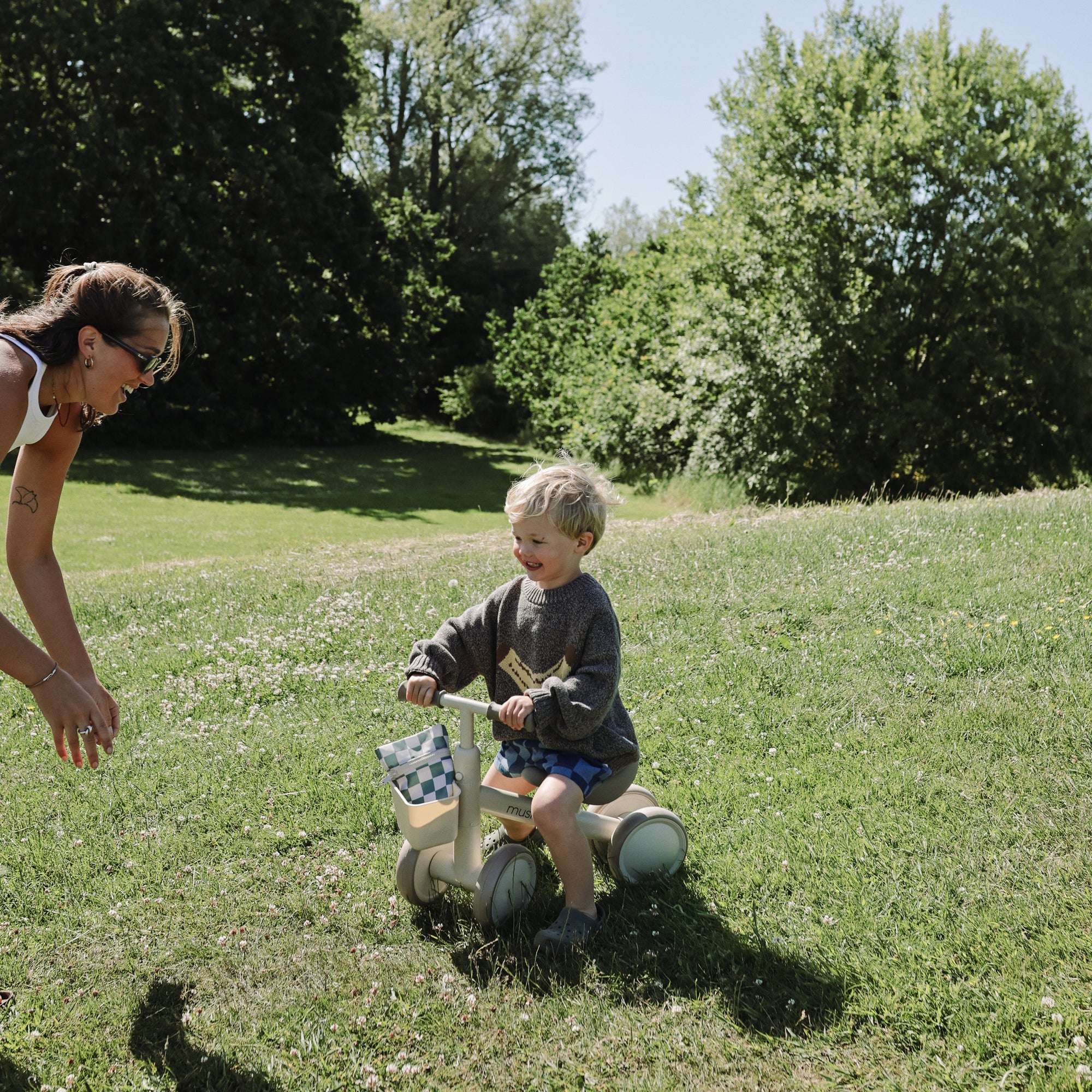 A child rides a mushie Mini Toddler Bike on grass as an adult reaches out on a sunny day.