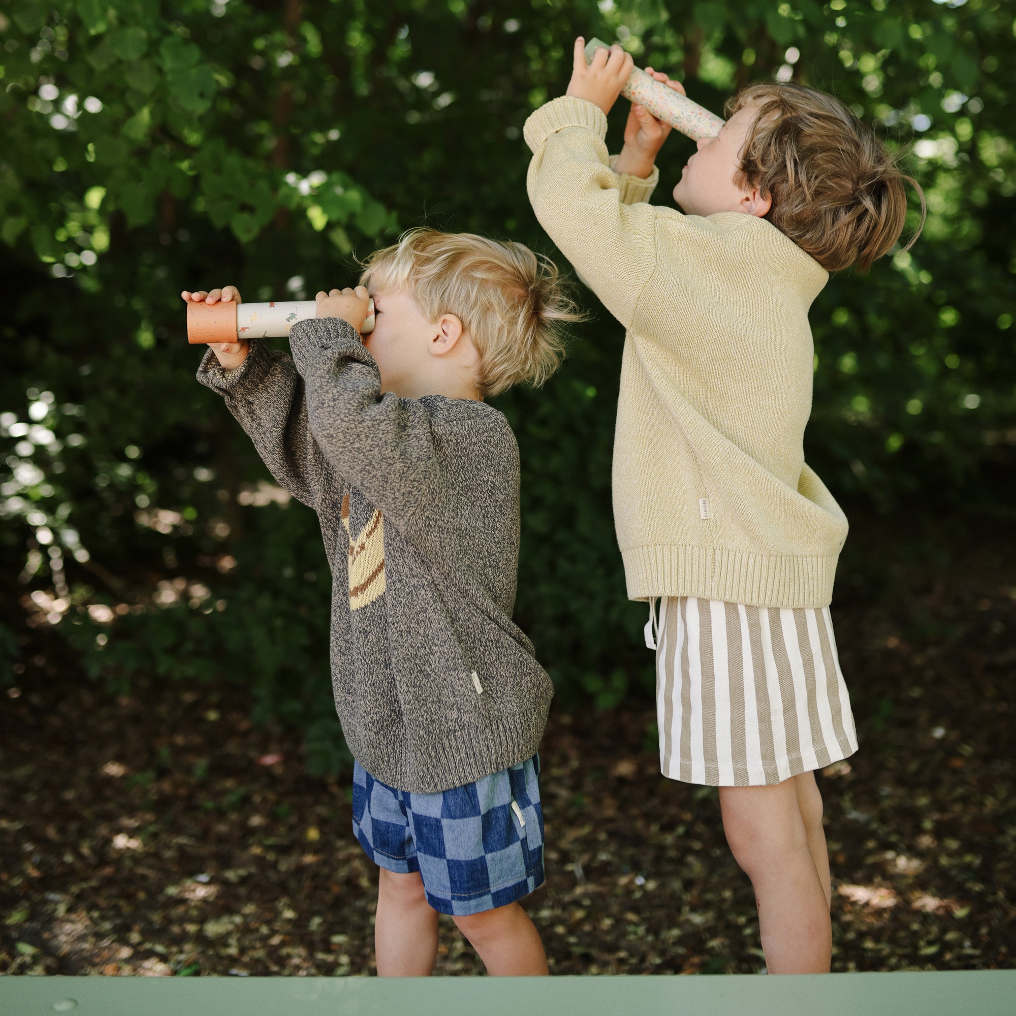 Two young children explore with mushie Kaleidoscopes surrounded by lush greenery.