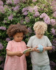 Two children hold mushie Nesting Spheres Sensory Toys, wearing striped summer outfits.