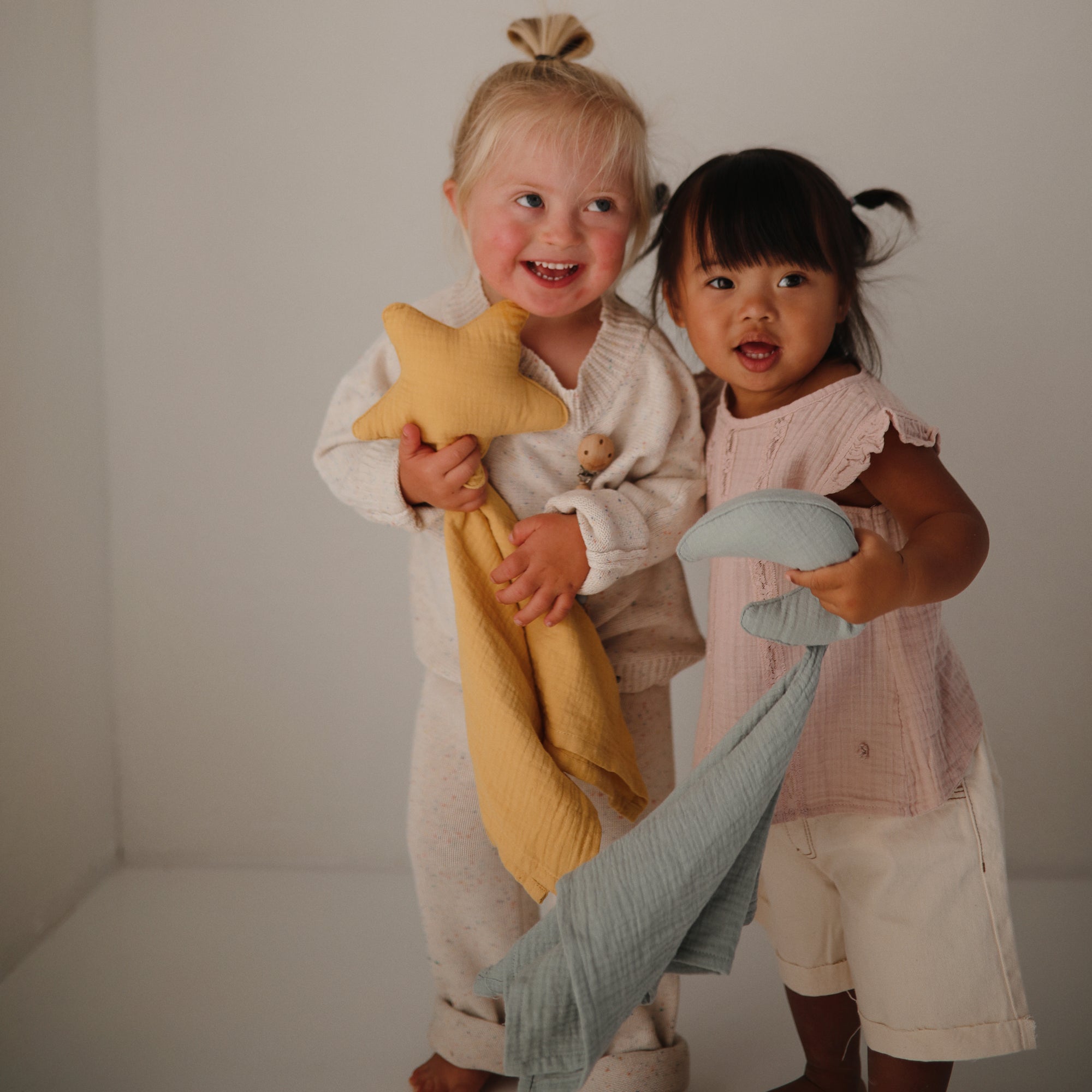 Two girls smile indoors, holding mushie Star Lovey Blankets and plush toys.