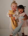 Two girls smile indoors, holding mushie Star Lovey Blankets and plush toys.