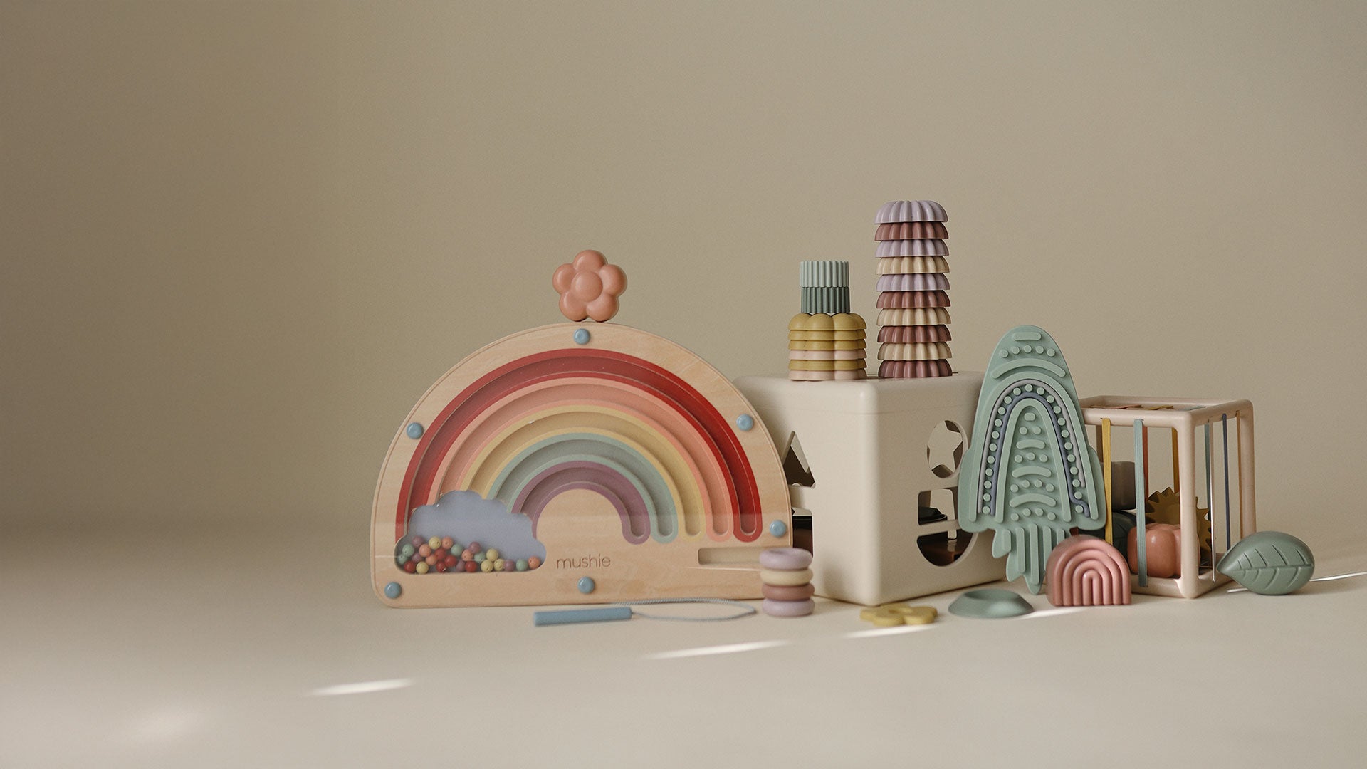 A set of pastel-colored wooden baby toys arranged on a warm beige studio background, including a rainbow-shaped toy, a white block with stacked patterned pieces, a blue rocket toy, an elastic box, and soft round accessories.