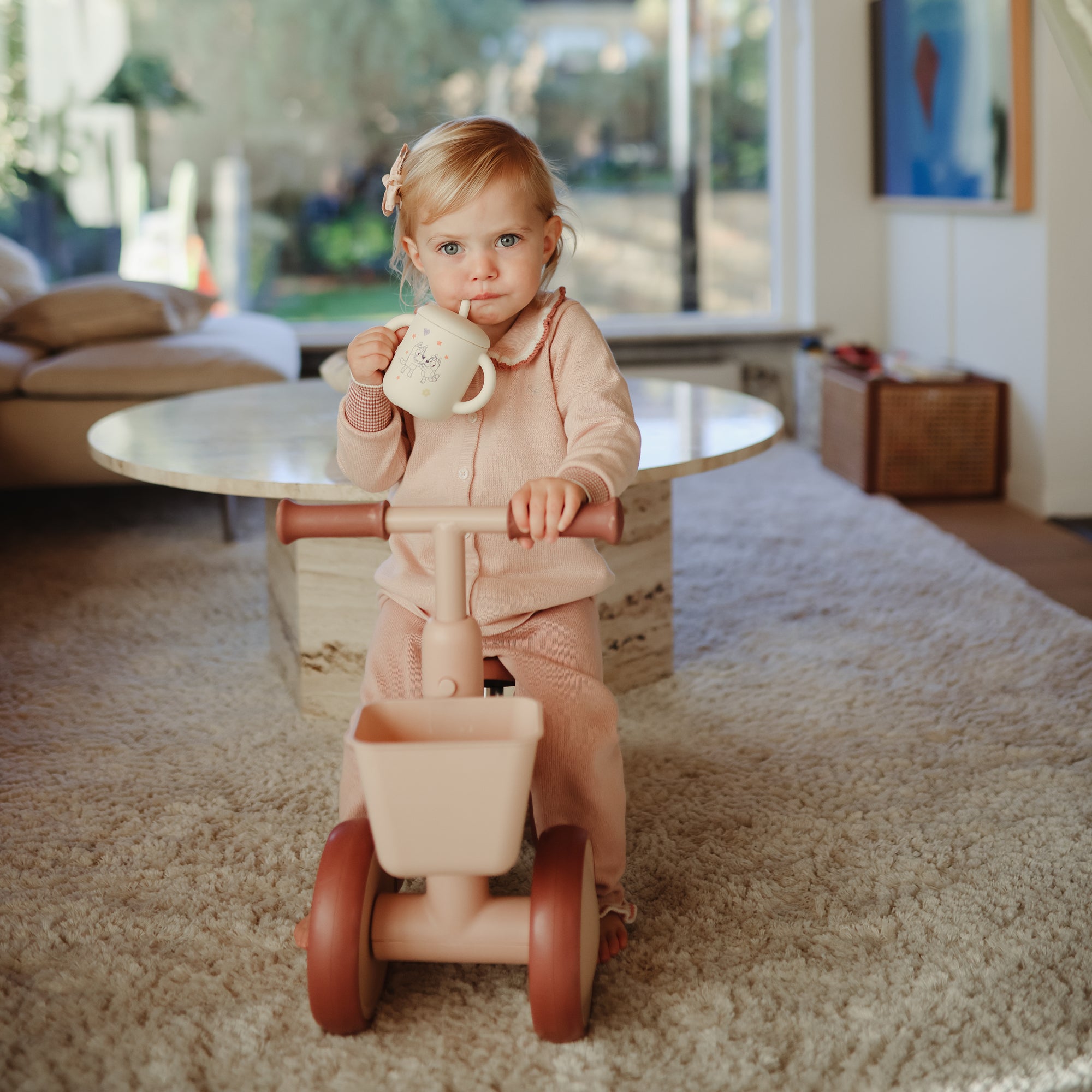 Toddler in pink pajamas on a trike holds a mushie Bluey Gift Set mug on a carpeted floor.
