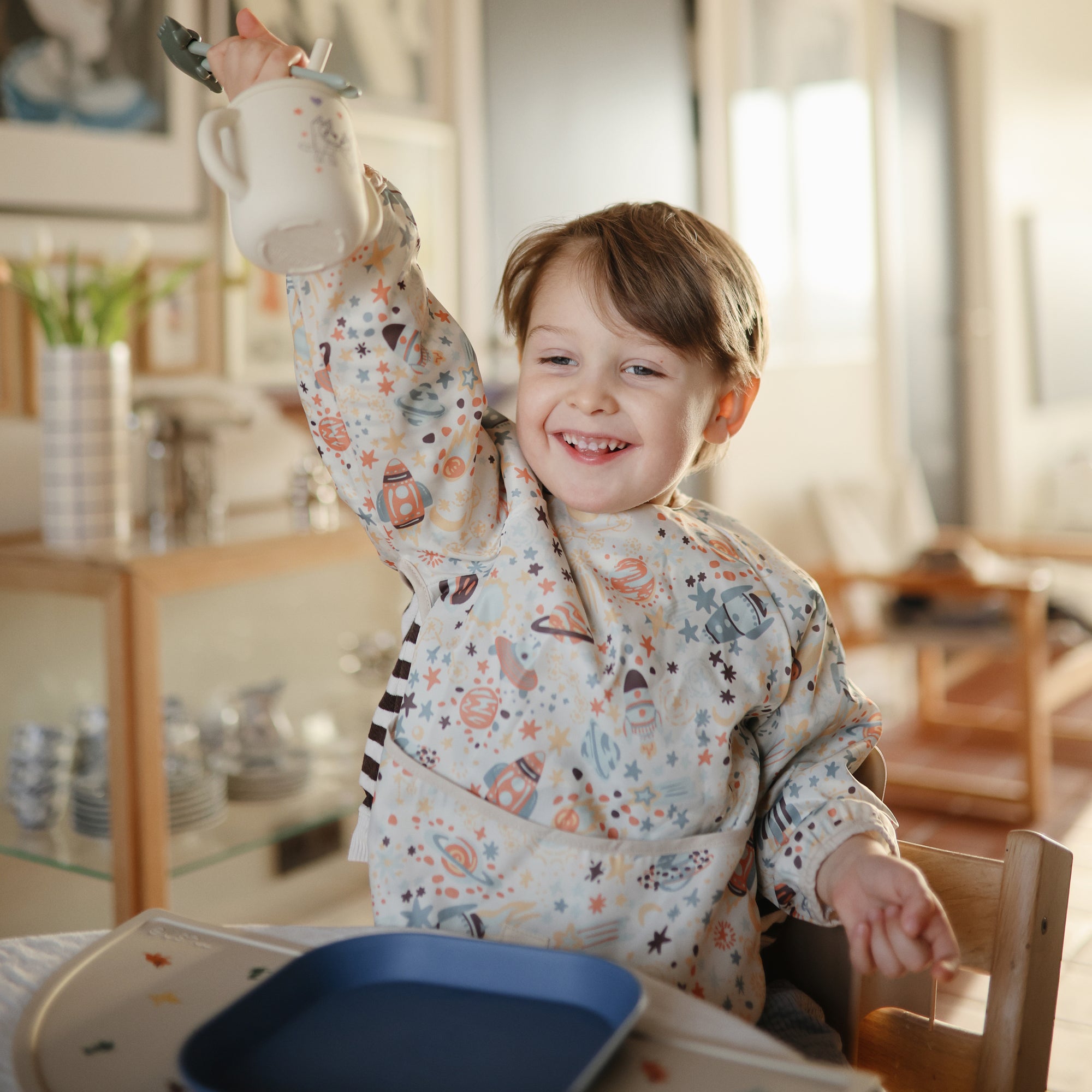 Smiling child in a Mushie Bluey Silicone Training Cup + Straw, seated at a table with a blue plate.