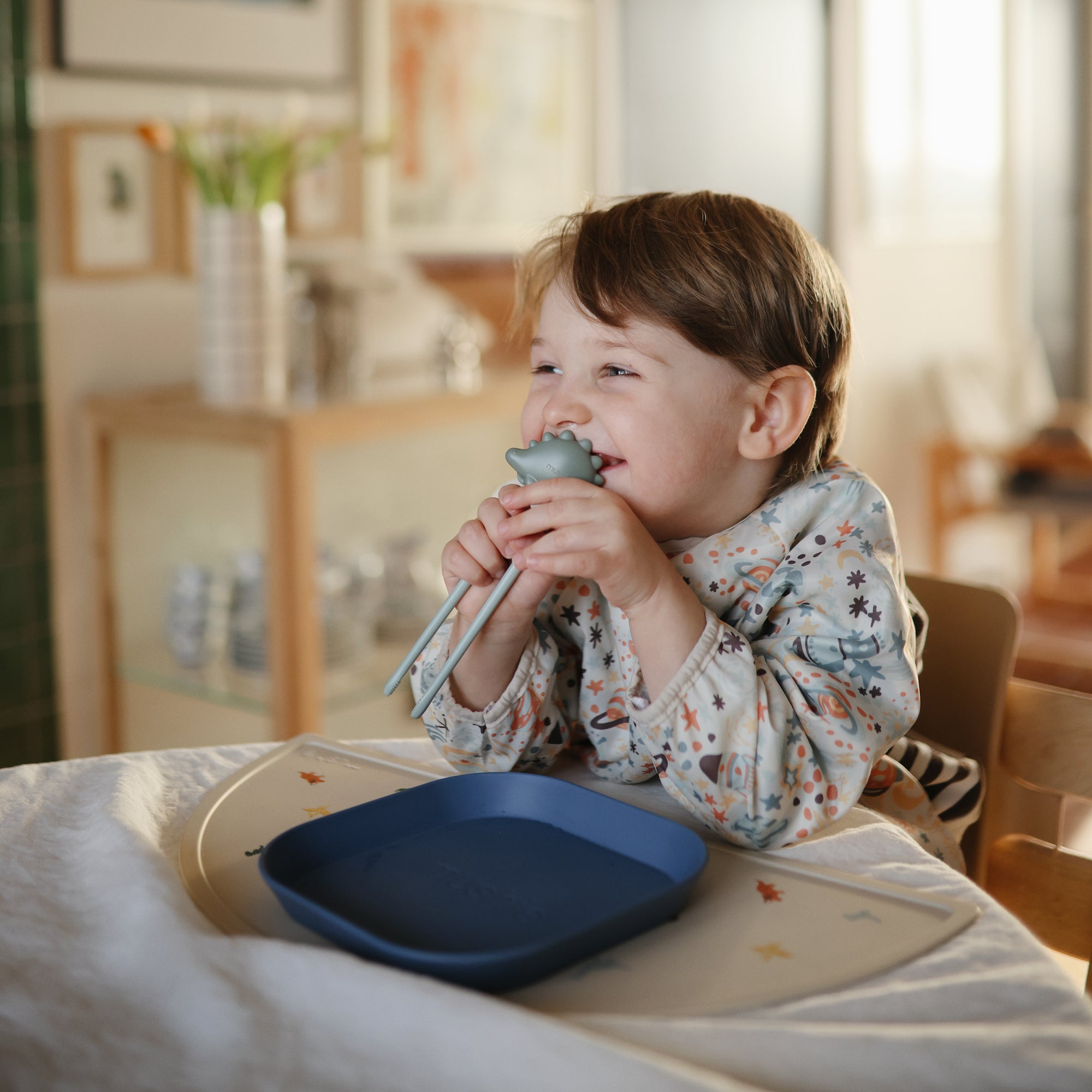 Smiling child in a mushie Long Sleeve Bib holds chopsticks at table with blue plate.