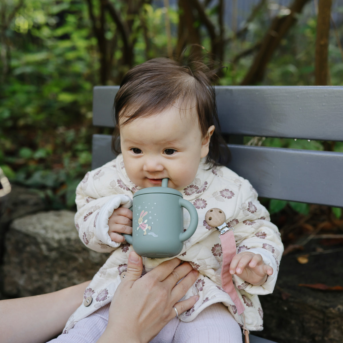 Baby on bench holding Mushie Silicone Training Cup + Straw, in patterned coat and pale leggings.