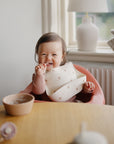 Smiling baby wears mushie Silicone Baby Bib in a highchair, holding food by a bowl.