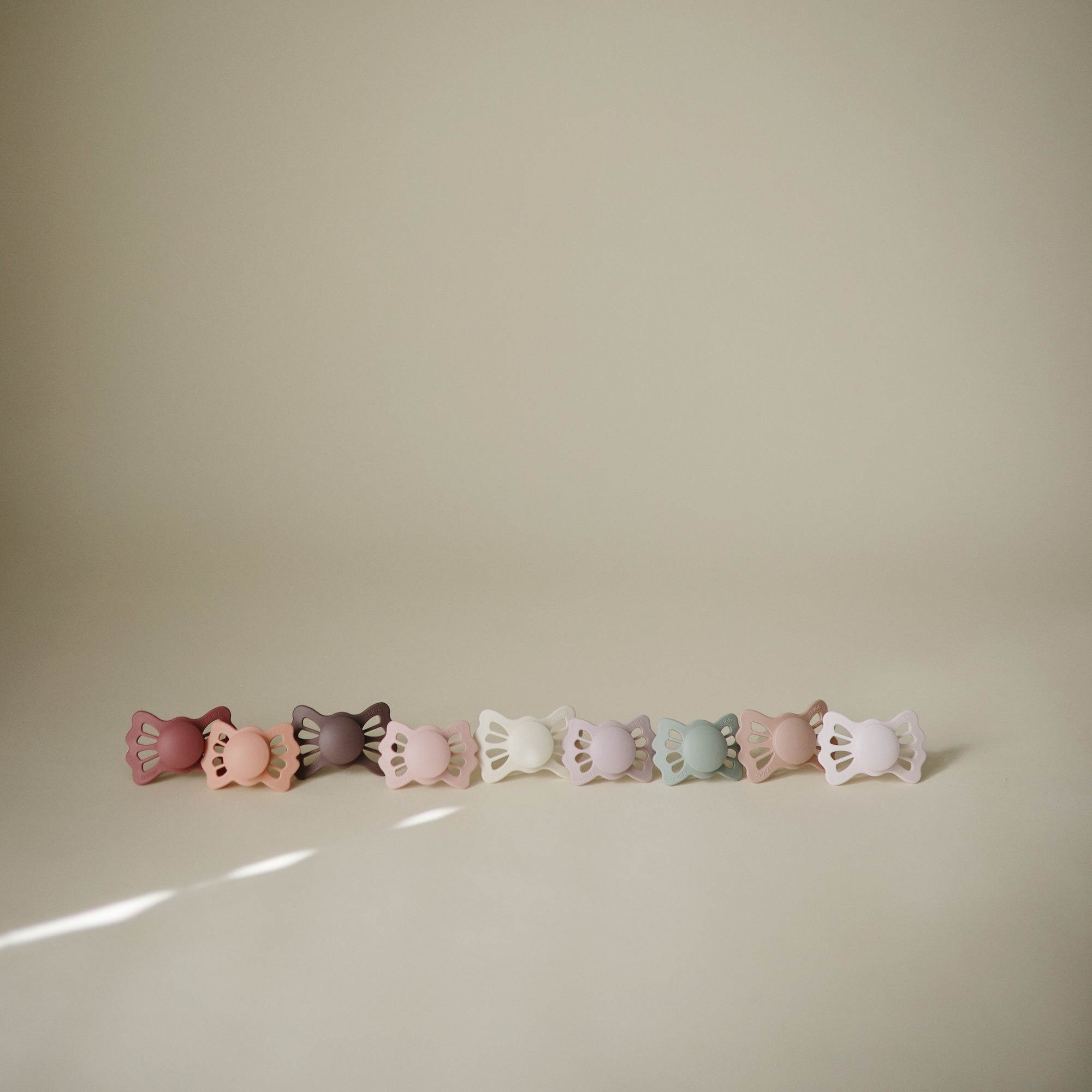 Eight FRIGG Lucky pacifiers, pastel-colored, are lined up against a beige background.