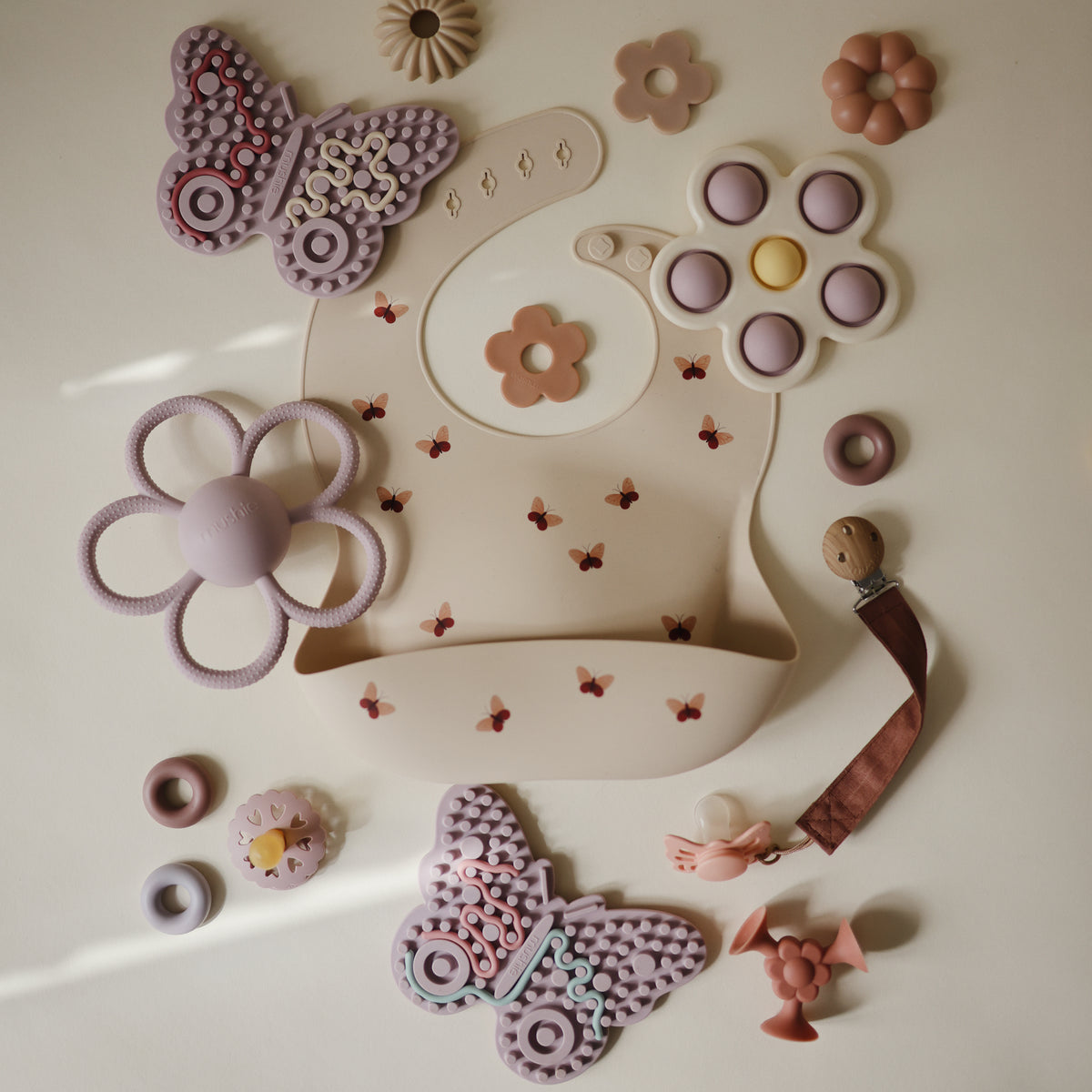 Mushie Silicone Sensory Toy and bibs in pink and purple on a light background.
