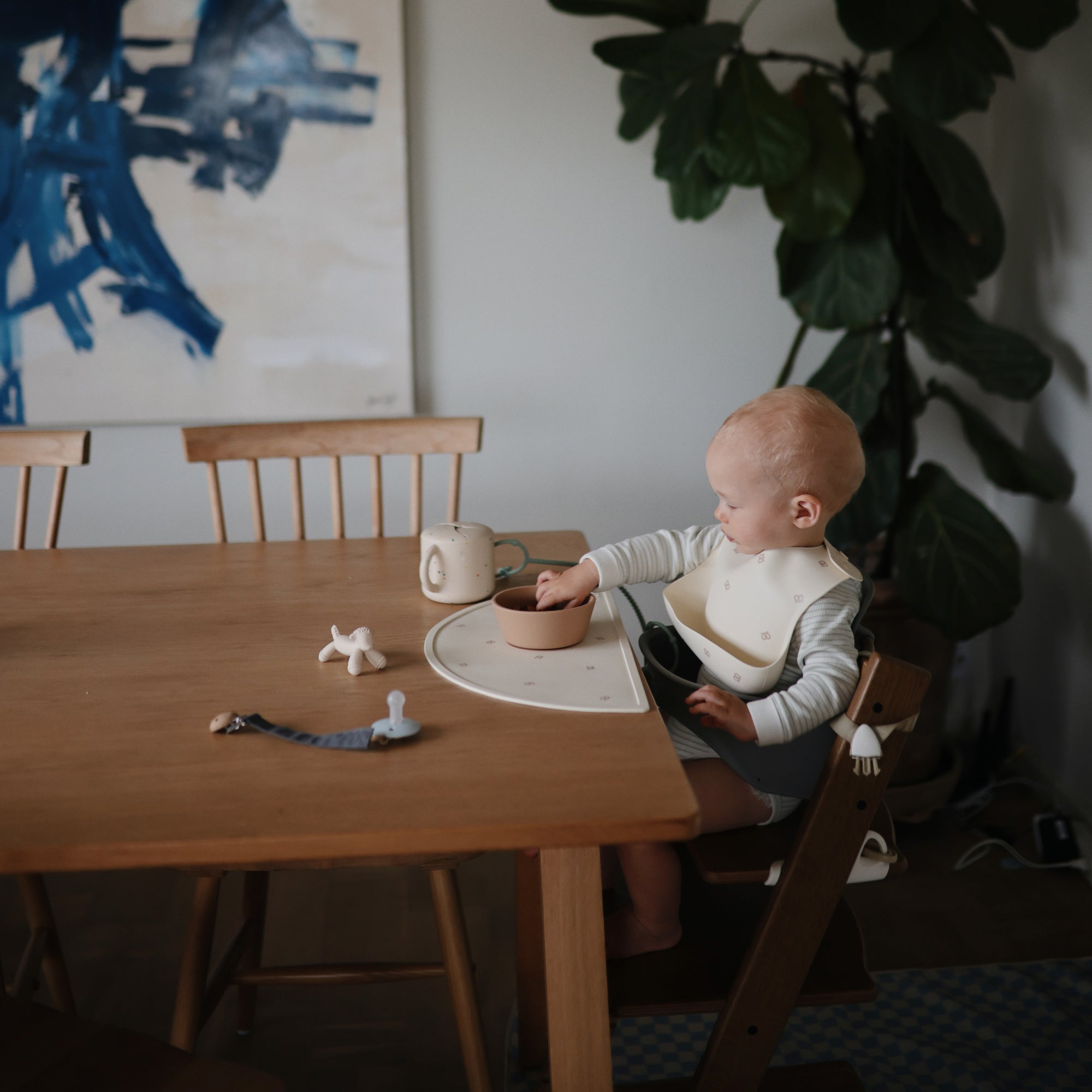 A baby with a mushie Figurine Teether sits at a wooden table in a cozy, modern dining area.