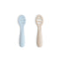 Two Mushie First Feeding Baby Spoons, one light blue and one beige, lie side by side on a white surface. These silicone teething spoons have a simple rounded design and cut-out slots in each spoon bowl.