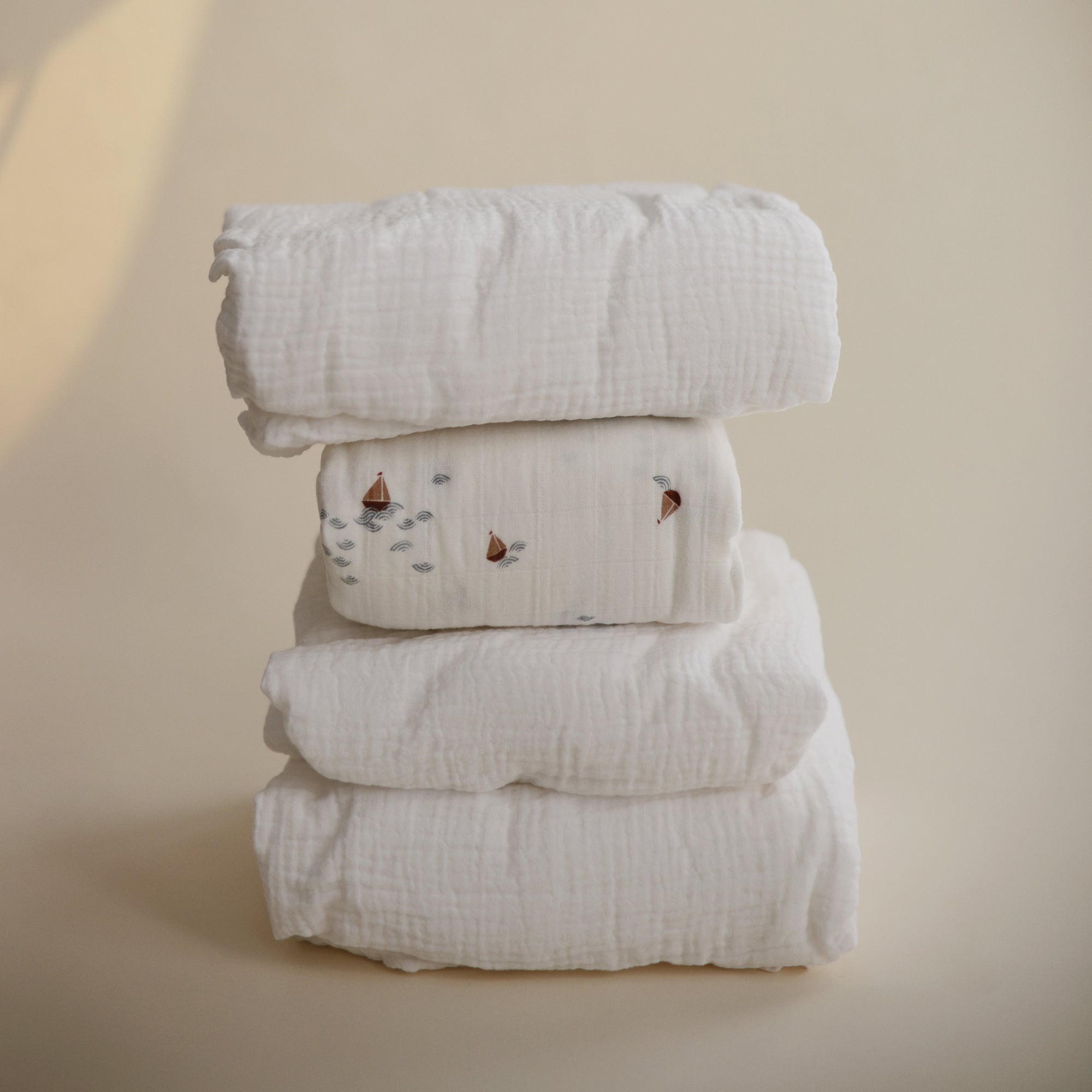 A stack of four mushie Extra Soft Muslin Crib Sheets, one with small sailboat prints.