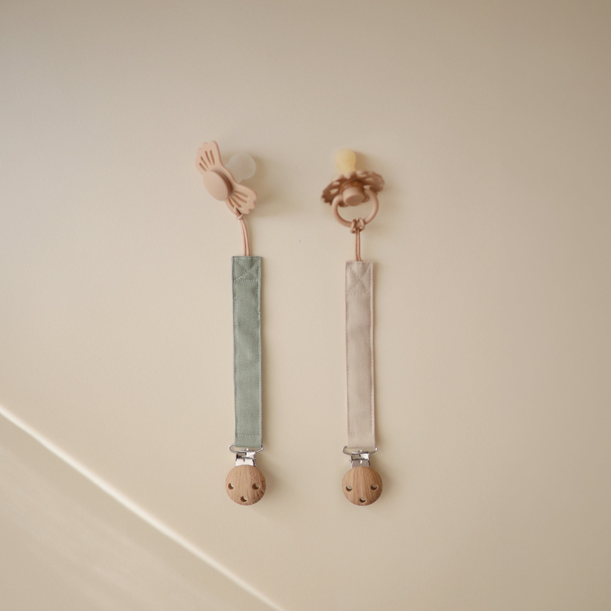 Two mushie Linen Pacifier Clip 2-Packs with wood ends hang on a beige wall.