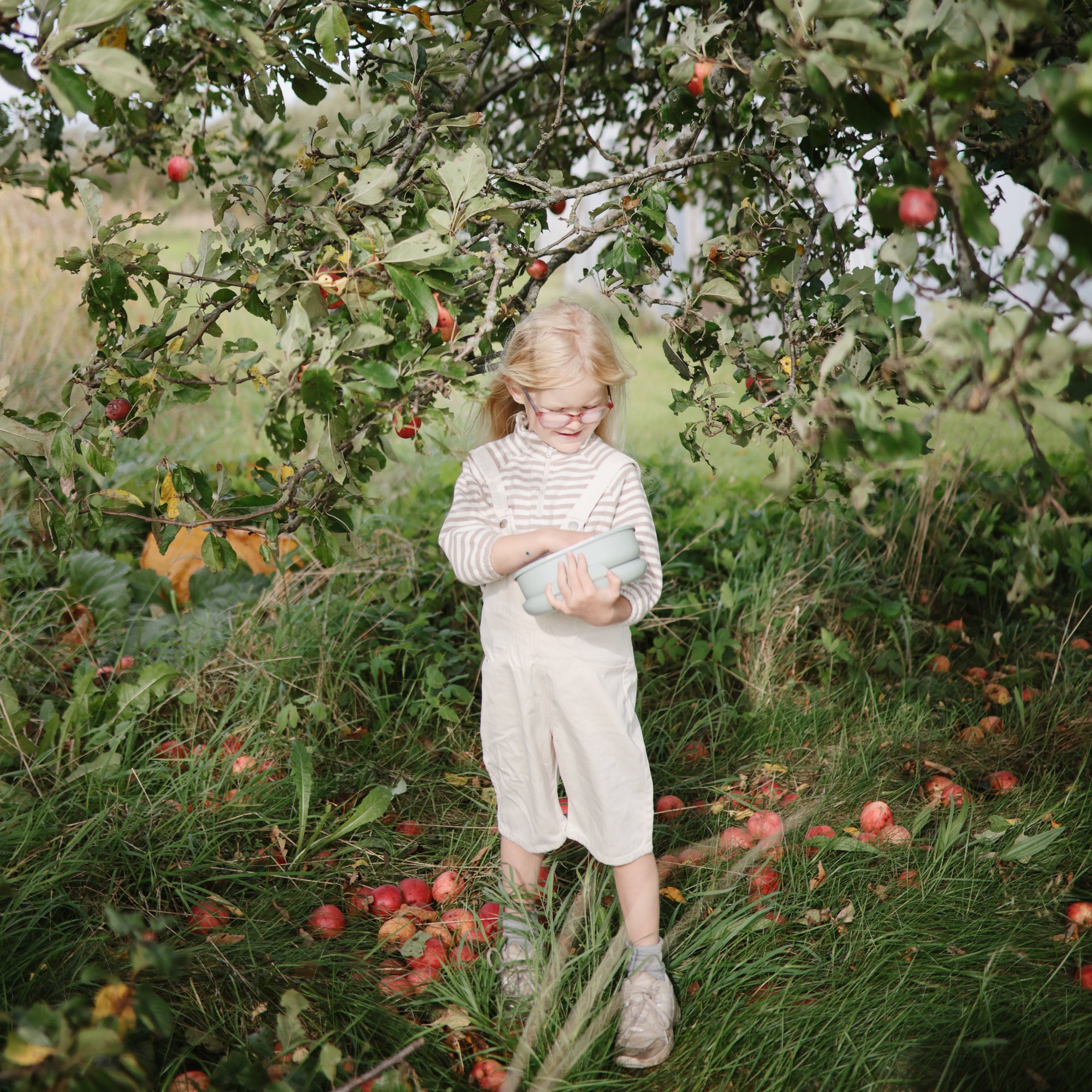 Smiling child uses mushie Lunchbox with compartments under an apple tree.