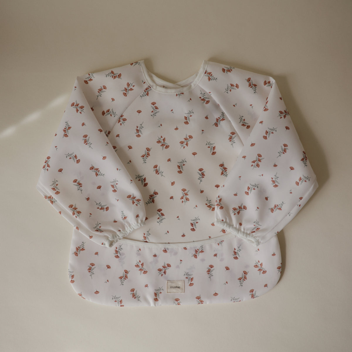 Mushie Long Sleeve Bib in a red floral print with a front pocket and wipe-clean fabric.