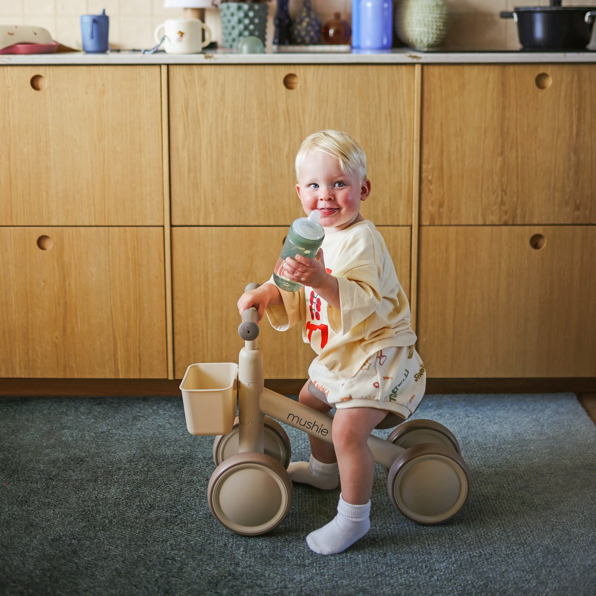 A toddler on a tricycle holds a mushie Soft Spout Sippy Cup in a cozy kitchen.