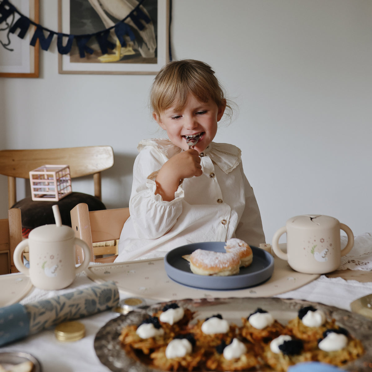 Smiling toddler enjoys a pastry, holding the mushie Snack Cup at the table.