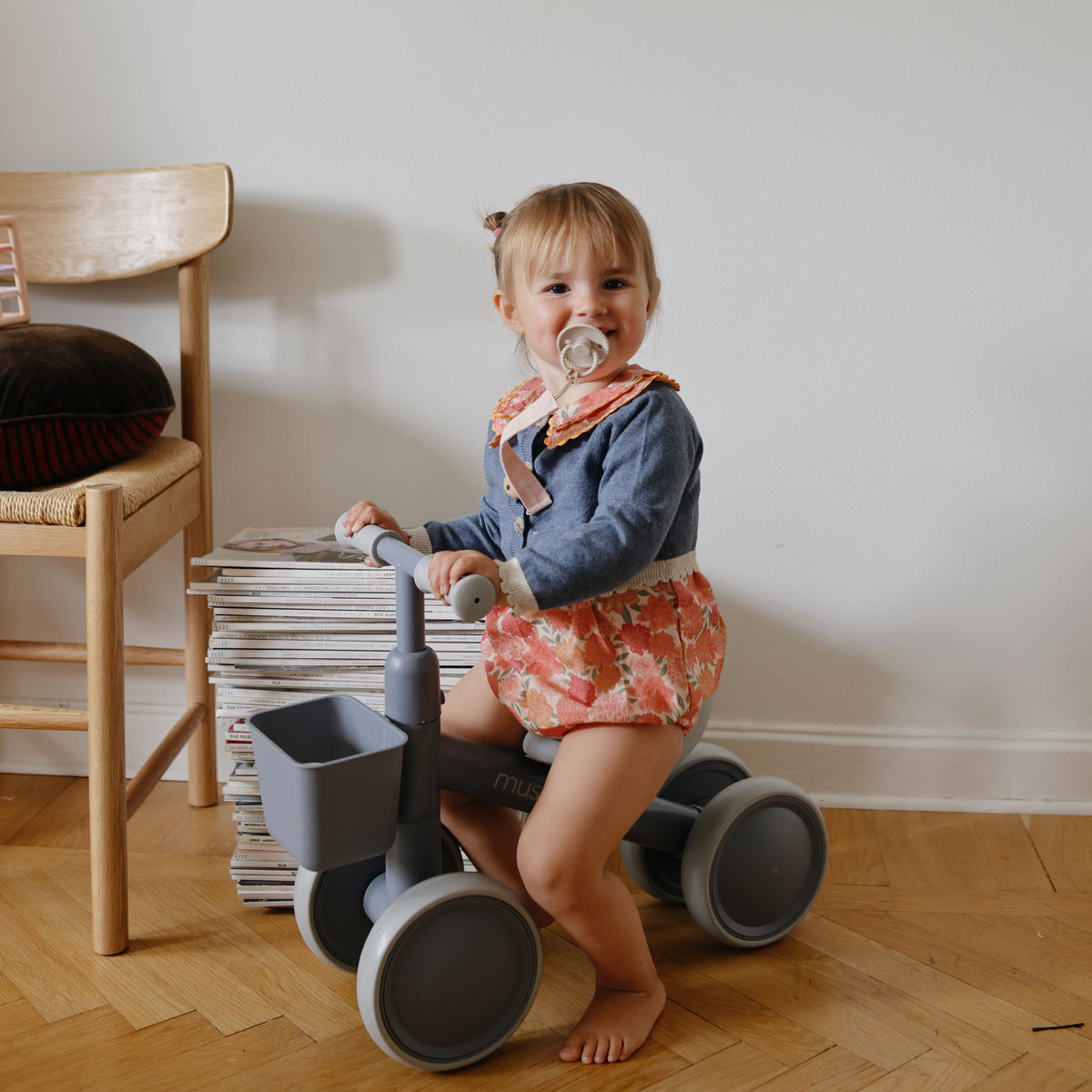 Toddler with pacifier sits on a mushie Mini Toddler Bike indoors next to a stack of magazines.