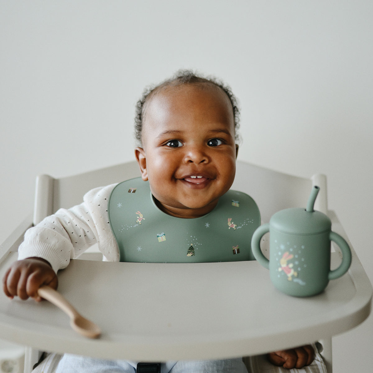 Smiling baby in a high chair wearing a green mushie Silicone Baby Bib, holding spoon and cup.