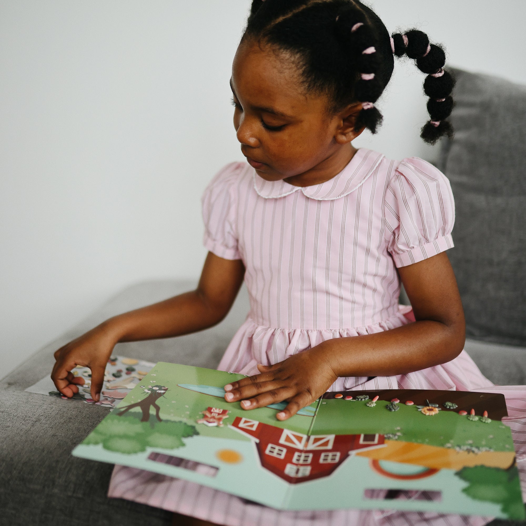 Young girl reads mushie Reusable Sticker Set on a gray sofa.
