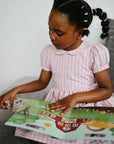 Young girl reads mushie Reusable Sticker Set on a gray sofa.