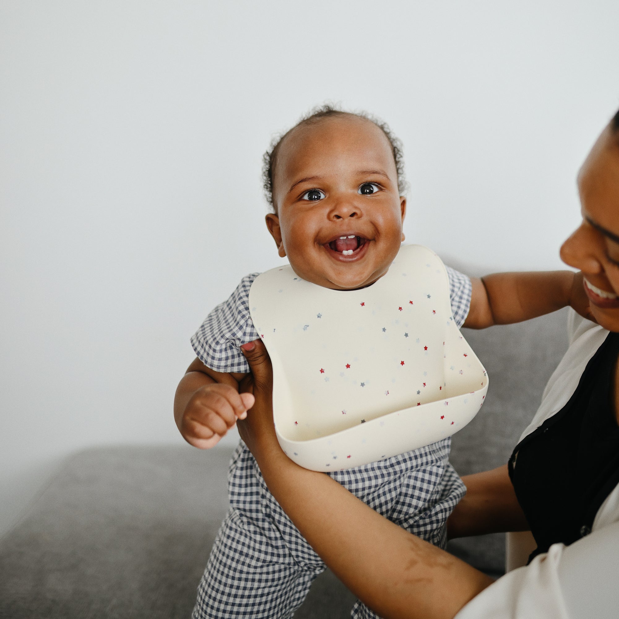 Smiling baby in mushie Silicone Baby Bib with adjustable neck strap, held by adult on gray couch.