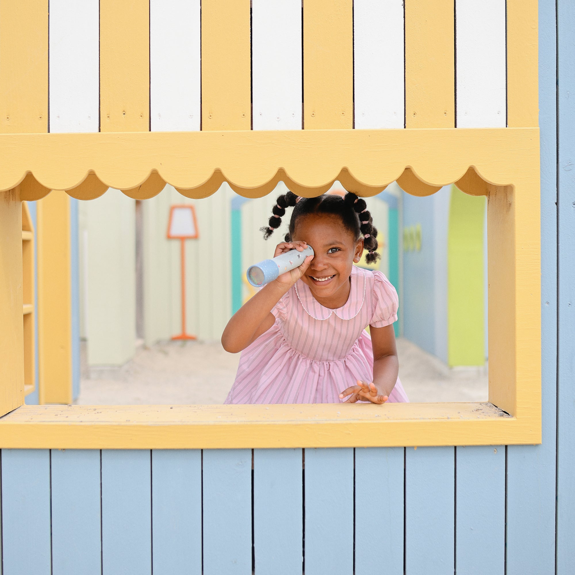 Girl in pink dress enjoys imaginative play with mushie Kaleidoscopes by the window.