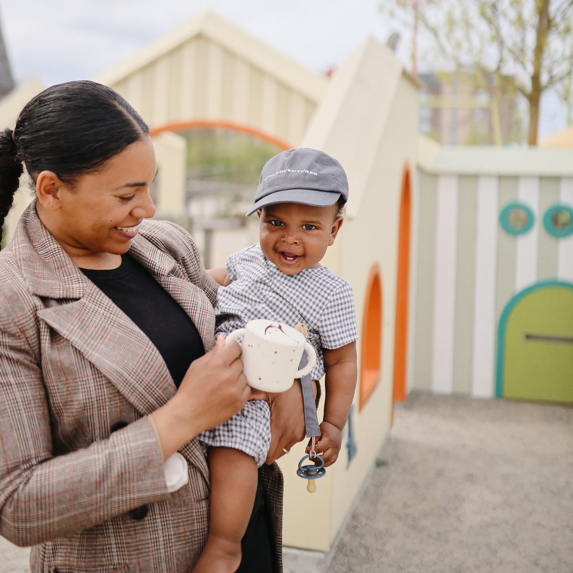 Smiling woman holds baby with FRIGG Moon Natural Rubber Pacifier 2-Pack at playground.