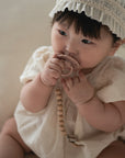 Baby in white with headband holds FRIGG Daisy Natural Rubber Pacifier 2-Pack with beaded clip.