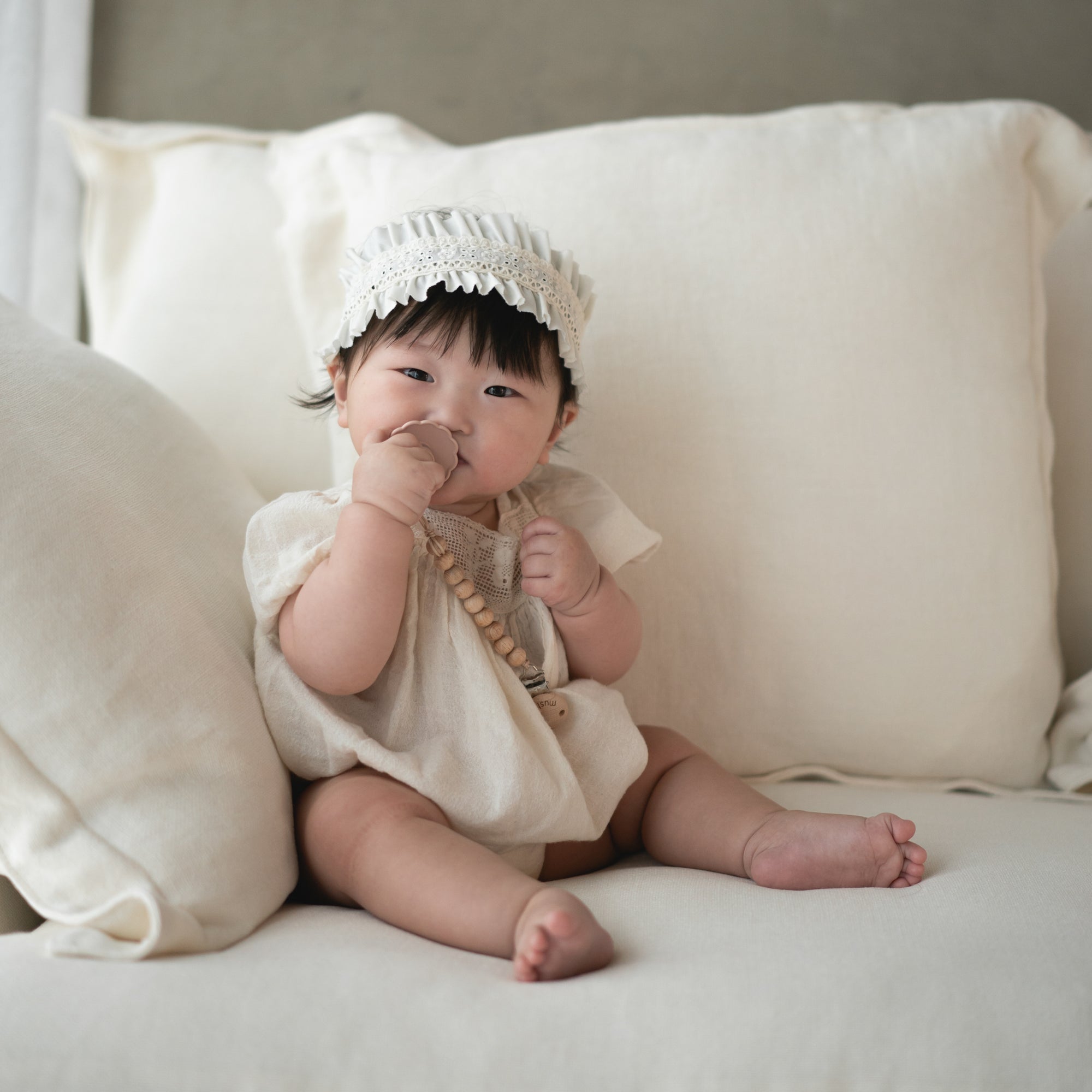 Baby in white holds FRIGG Daisy Natural Rubber Pacifier 2-Pack on a cream sofa.