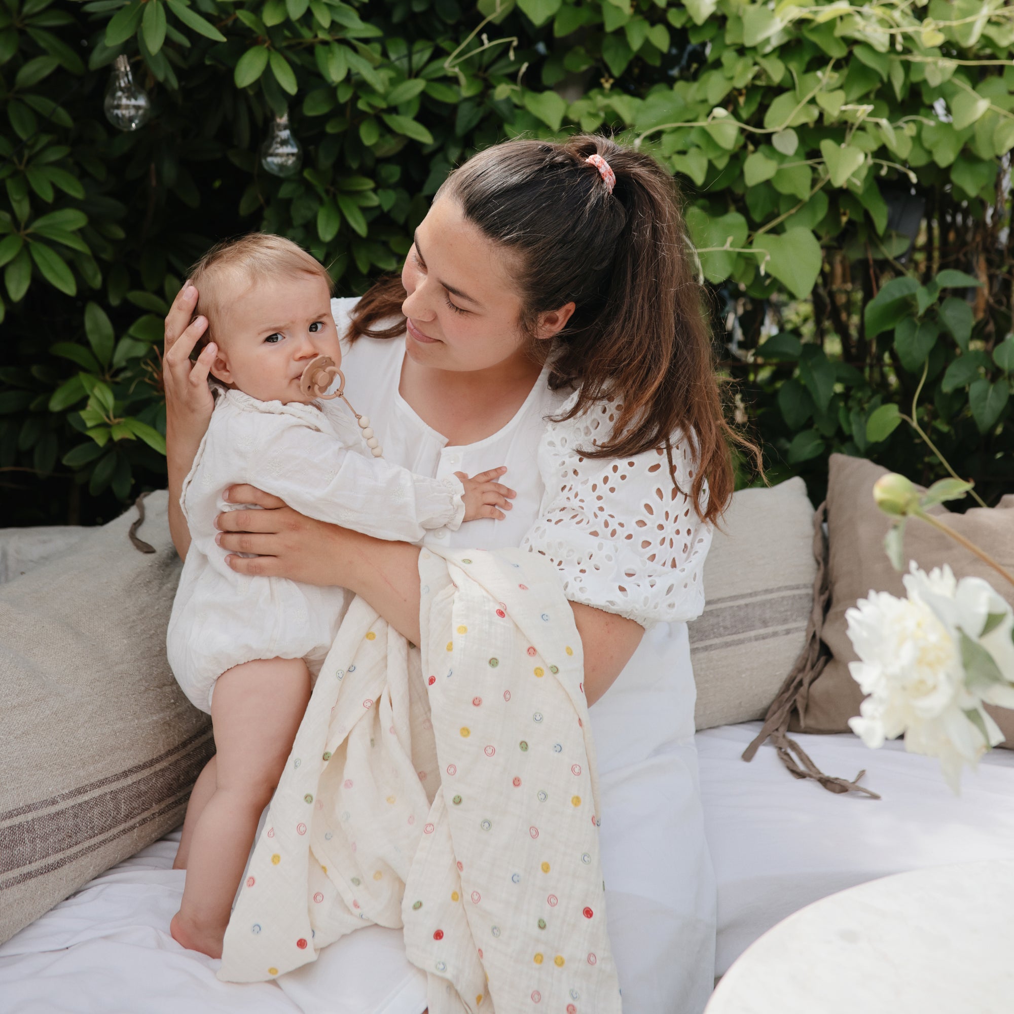 A woman in white holds a baby with a FRIGG Daisy Natural Rubber Pacifier near green foliage.