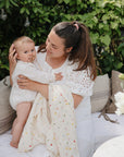 A woman in white holds a baby with a FRIGG Daisy Natural Rubber Pacifier near green foliage.