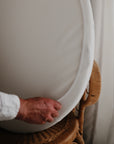 A hand places the mushie Stretchy MINI Crib Sheet, made from breathable organic cotton, onto a woven rattan chair beside a sheer white curtain, creating a cozy, inviting space.