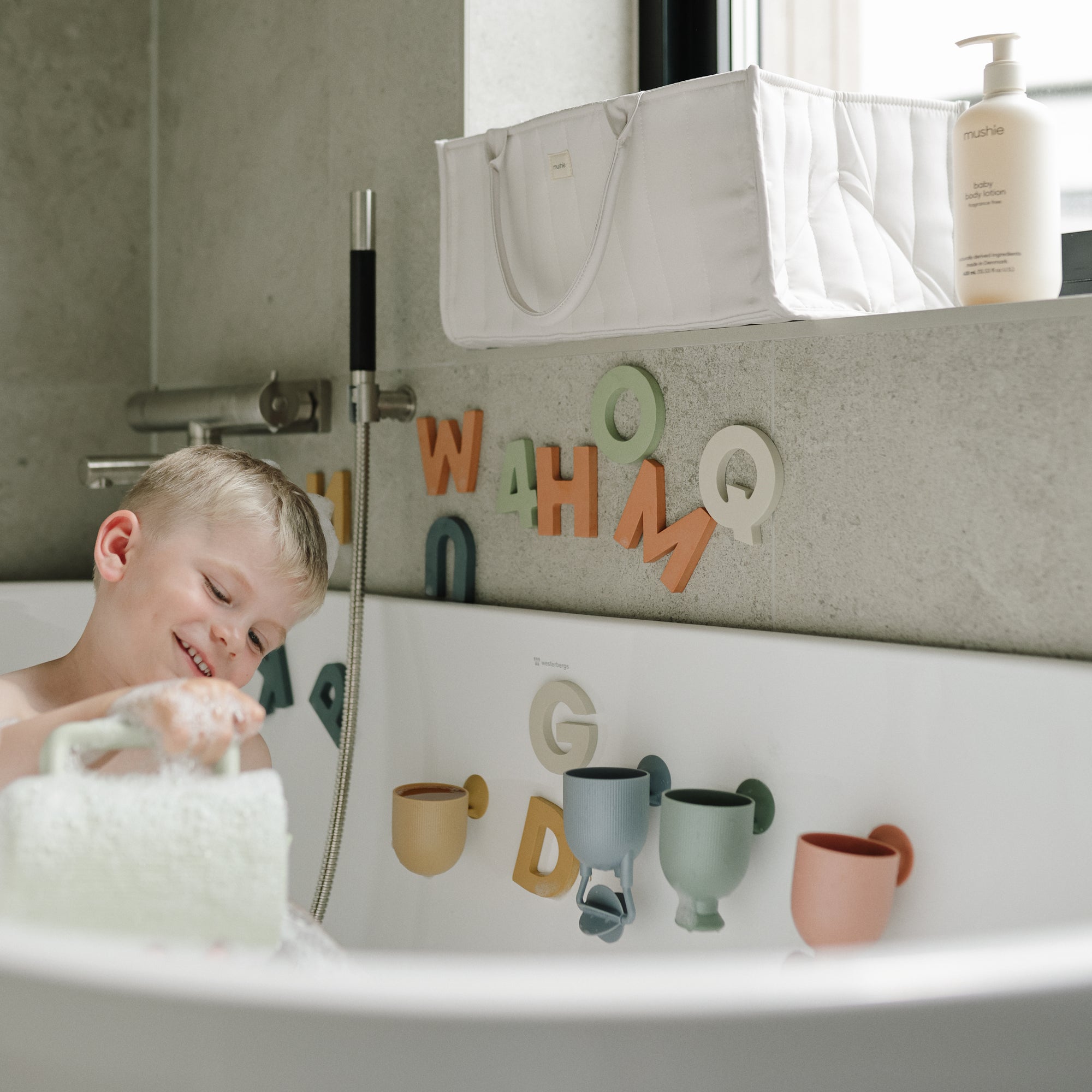 Smiling child plays in bathtub with mushie Letters &amp; Numbers Bath Play Set on the wall.