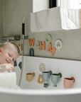 Smiling child plays in bathtub with mushie Letters & Numbers Bath Play Set on the wall.