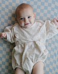 Smiling baby in a romper on mushie Extra Soft Muslin Crib Sheet with blue and white checks.