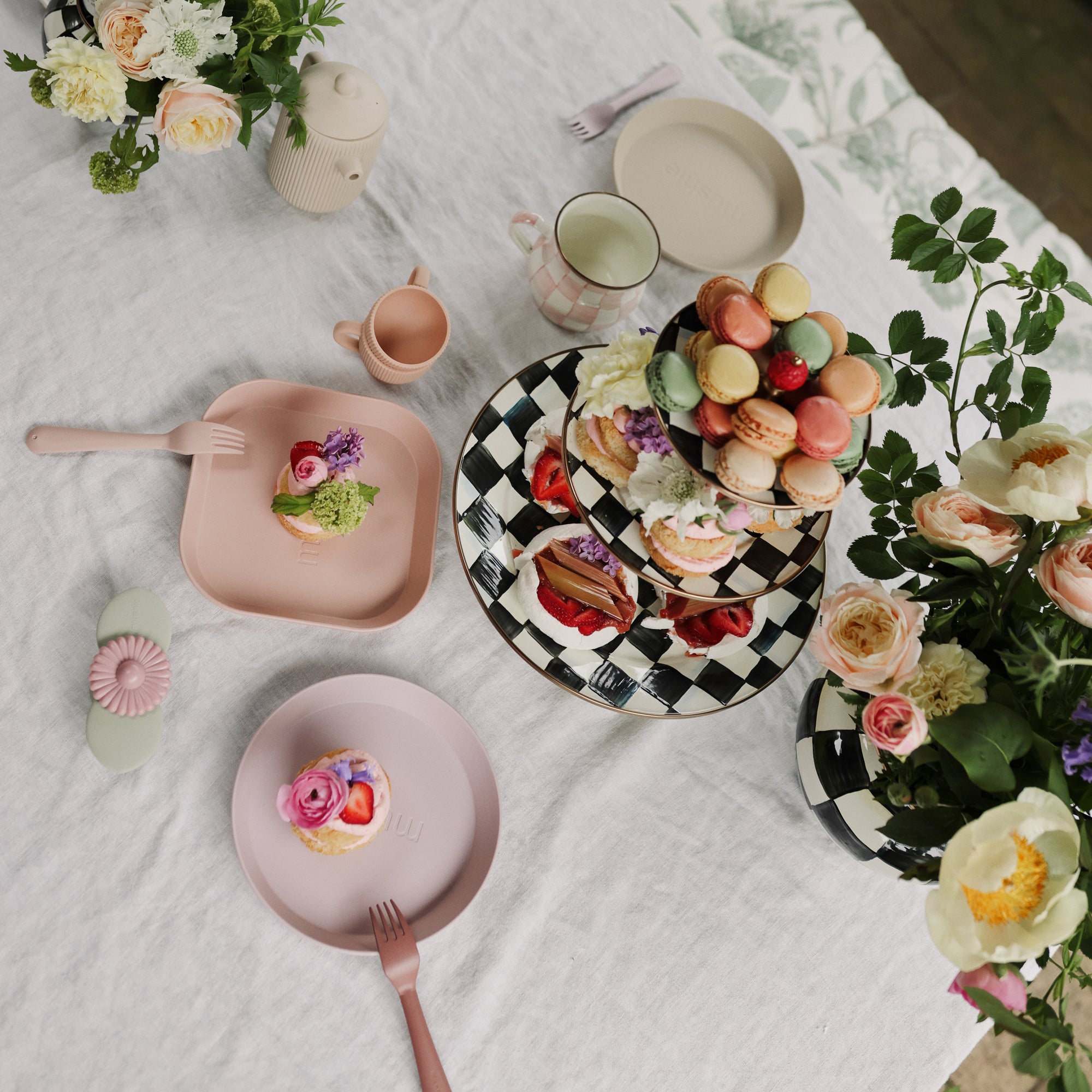 Mushie Round Dinnerware Plates, forks, pastries, macarons, and flowers on a white tablecloth.