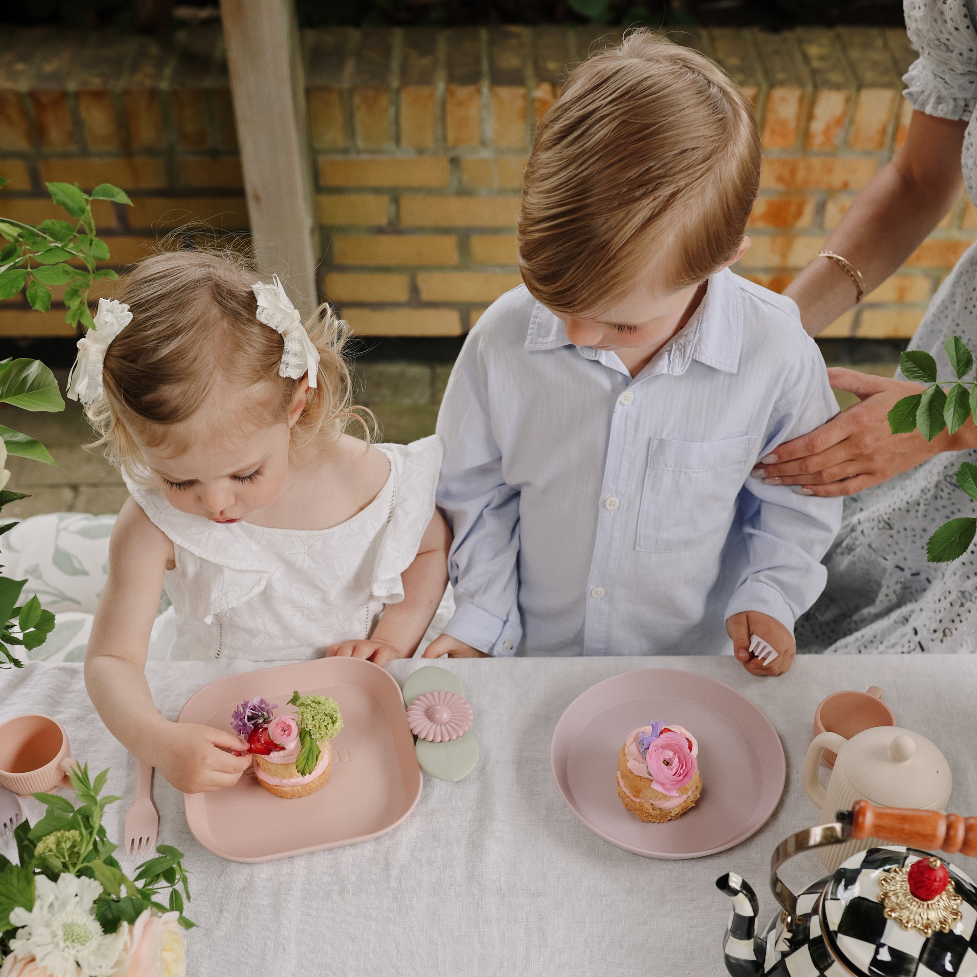 Two kids sit at a table with cakes on mushie Round Dinnerware Plates with pink flowers.