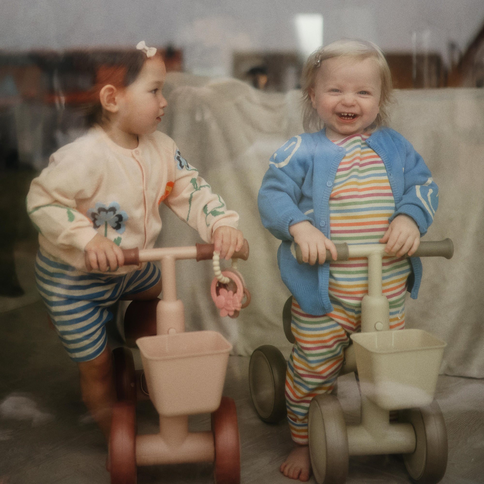Two toddlers indoors on toy bikes, each holding a mushie Fairy Teething Ring.