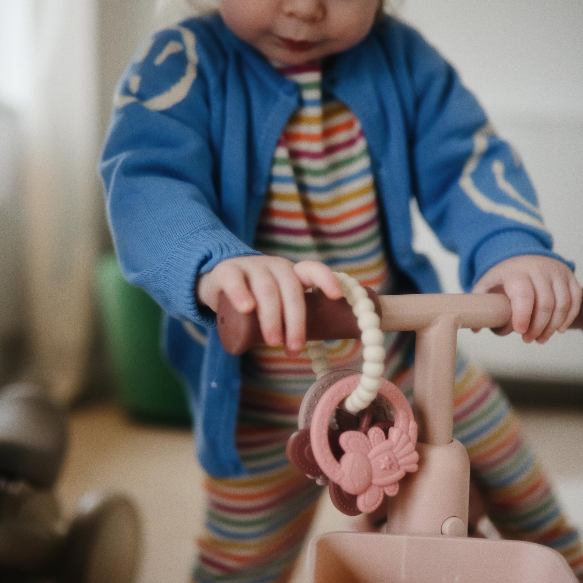 Toddler with mushie Fairy Teething Ring explores pink tricycle in striped outfit.