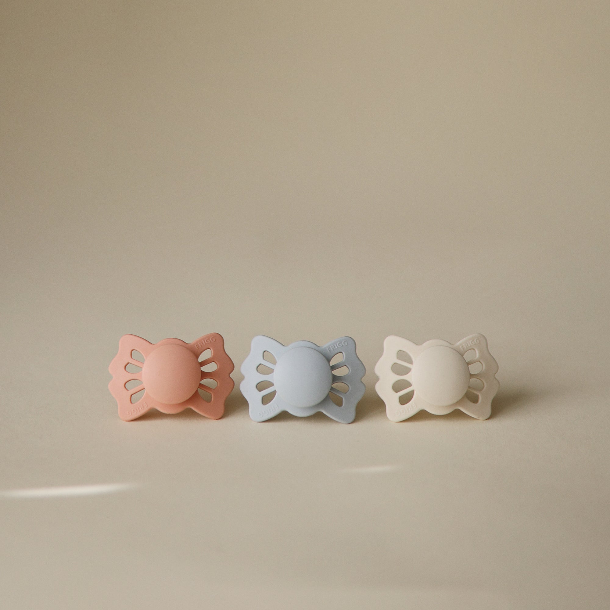 FRIGG Lucky Symmetrical Silicone Pacifier 2-Pack for 6-18 months in pink, blue, and beige.