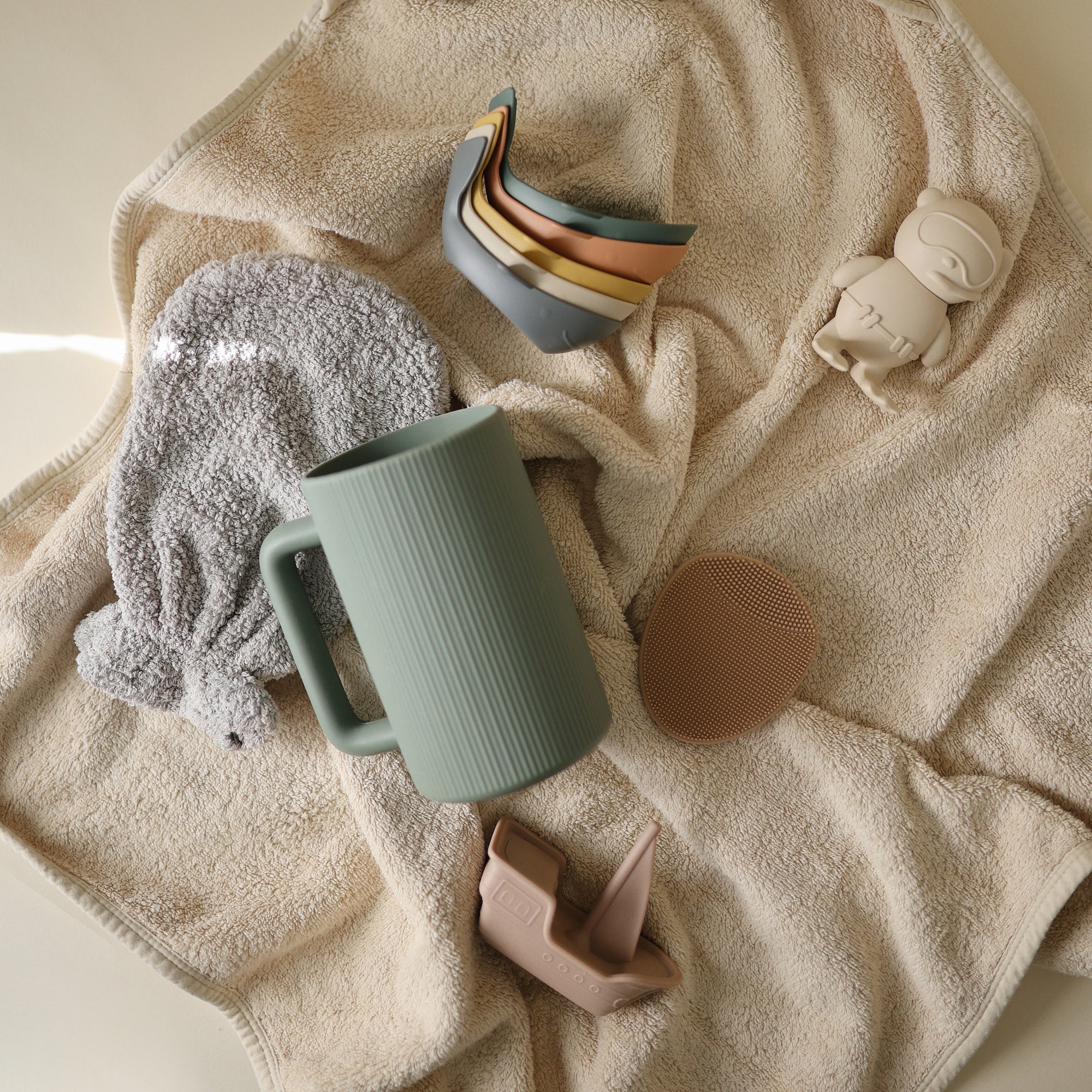 Mushie Whale Bath Stackers, sippy cups, and bibs on a beige towel.