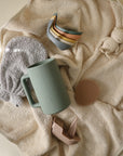Mushie Whale Bath Stackers, sippy cups, and bibs on a beige towel.