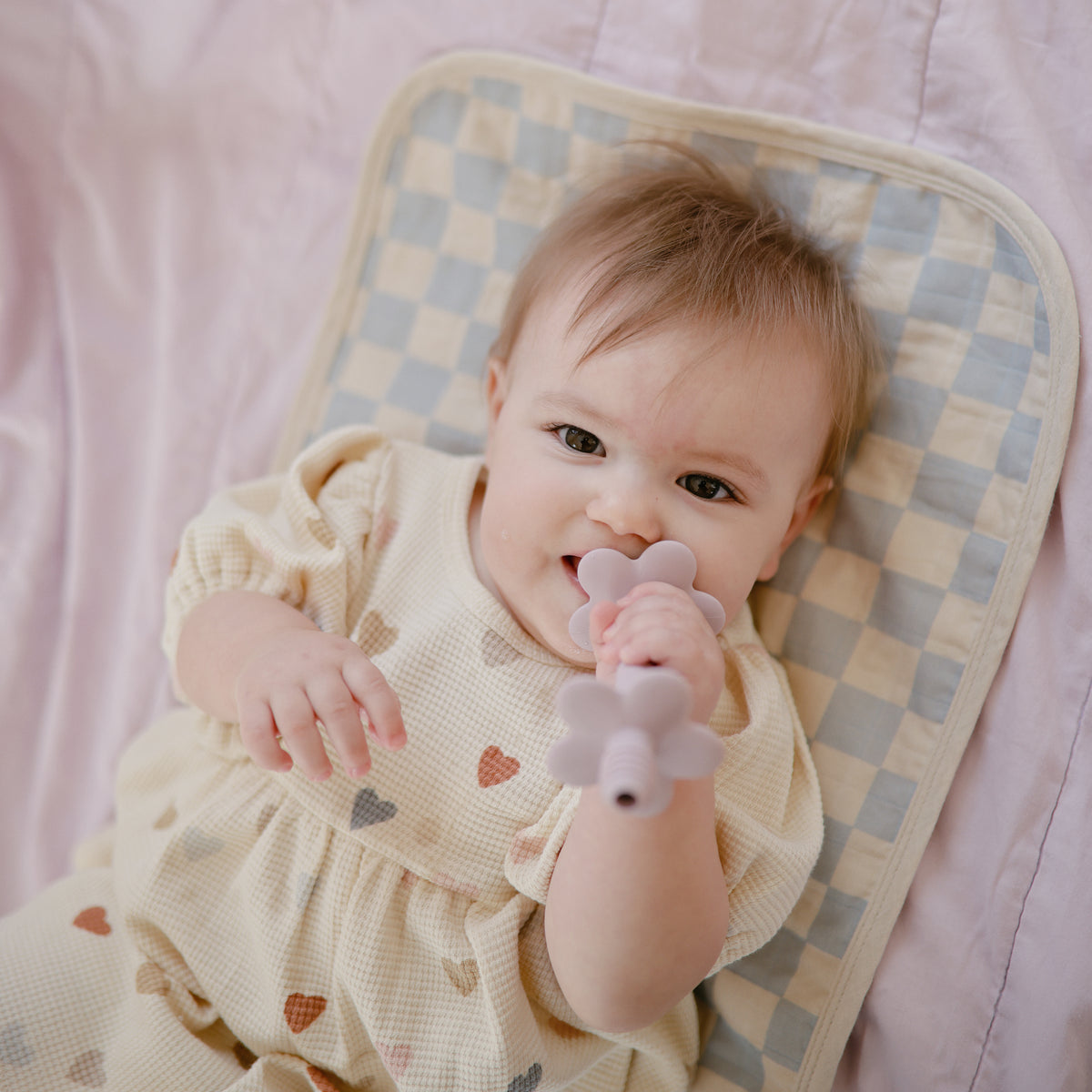 Baby chewing a flower teether on a Mushie Changing Pad Liner with a checkerboard pattern.