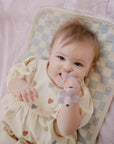 Baby chewing a flower teether on a Mushie Changing Pad Liner with a checkerboard pattern.
