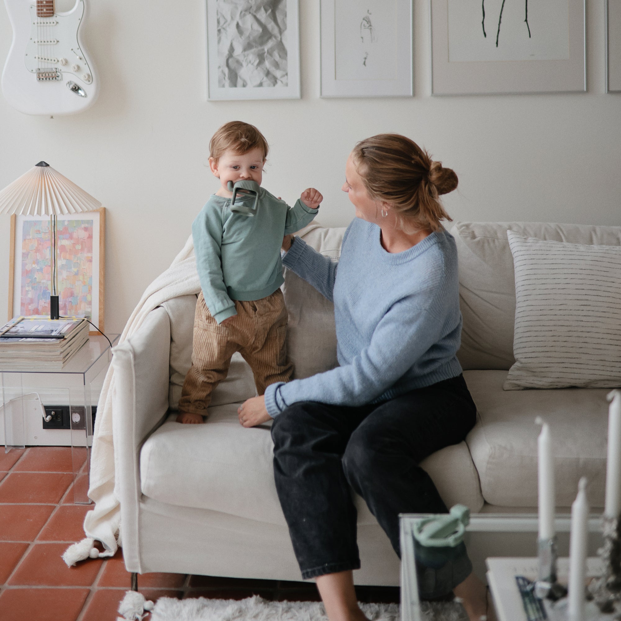 A woman smiles as a toddler explores the mushie No-Drop Teether on the couch indoors.