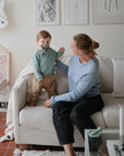 A woman smiles as a toddler explores the mushie No-Drop Teether on the couch indoors.